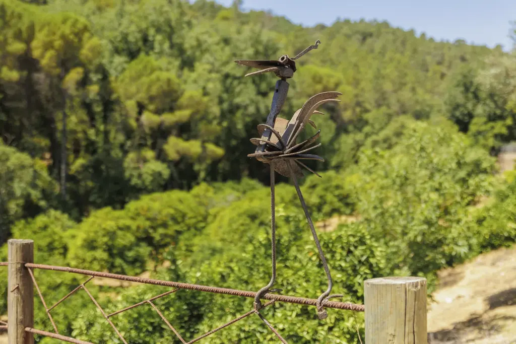Escultura de hierro de un pájaro sobre la valla, con el bosque de fondo